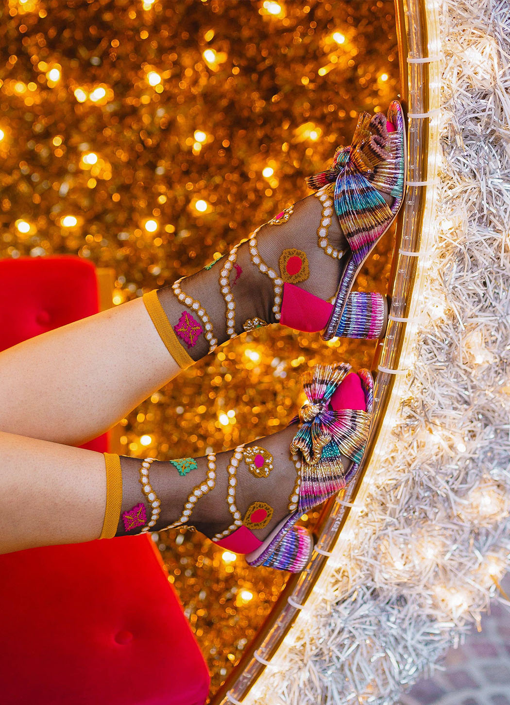 A person's legs wearing colorful, highly embellished high-heeled sandals with sheer patterned mesh and bright pink straps, posed against a glittering gold backdrop with a red chair visible on the left.