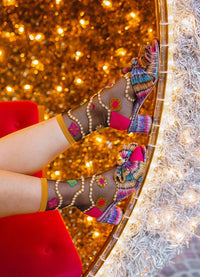 A person's legs wearing colorful, highly embellished high-heeled sandals with sheer patterned mesh and bright pink straps, posed against a glittering gold backdrop with a red chair visible on the left.