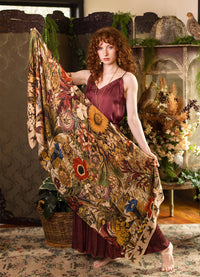 A woman with curly red hair stands barefoot in a rustic indoor setting, holding up a large patterned textile. The fabric features a dense floral and botanical print with sunflowers, roses, leaves, and birds in warm earthy tones of beige, brown, yellow, red, and green.