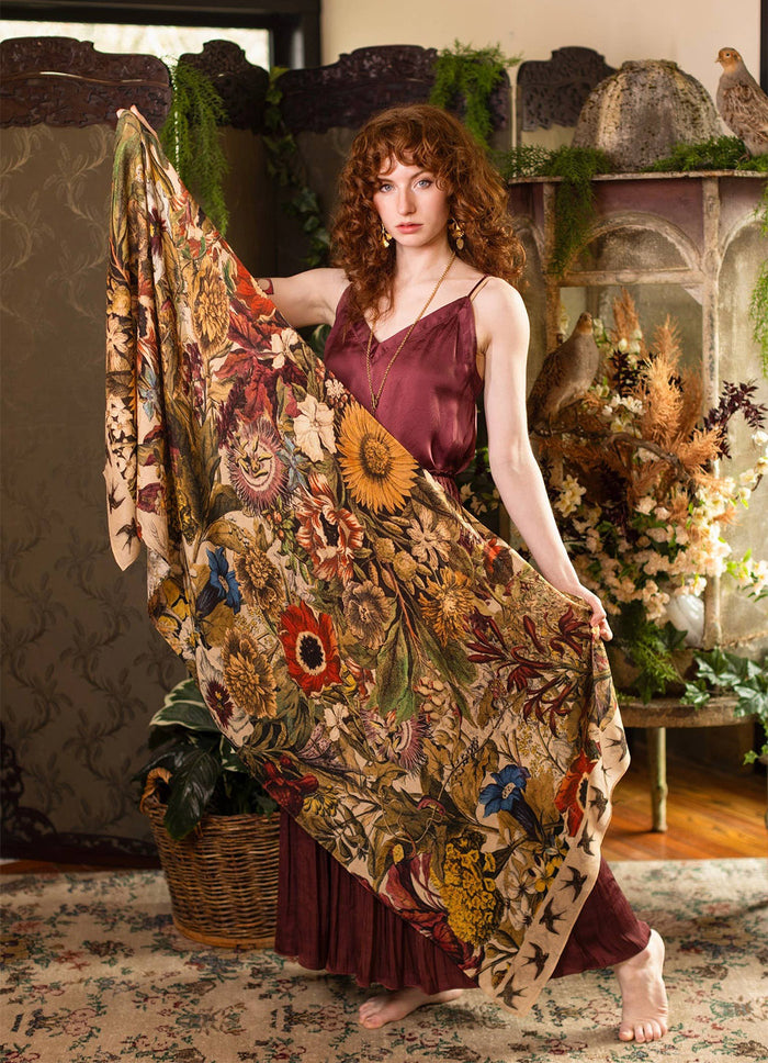 A woman with curly red hair stands barefoot in a rustic indoor setting, holding up a large patterned textile. The fabric features a dense floral and botanical print with sunflowers, roses, leaves, and birds in warm earthy tones of beige, brown, yellow, red, and green.