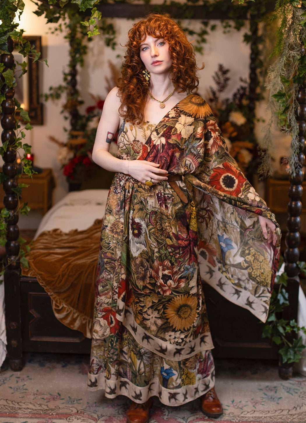 A woman with curly red hair stands in a decorated bedroom wearing a long bohemian floral print maxi dress with wide draped sleeves. The dress features a complex multicolor pattern of flowers, sunflowers, and butterflies on a beige background.