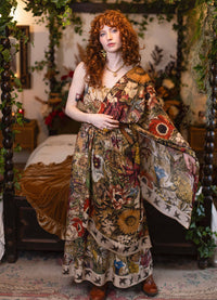 A woman with curly red hair stands in a decorated bedroom wearing a long bohemian floral print maxi dress with wide draped sleeves. The dress features a complex multicolor pattern of flowers, sunflowers, and butterflies on a beige background.
