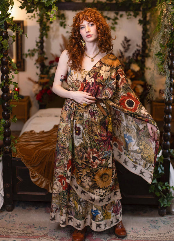 A woman with curly red hair stands in a decorated bedroom wearing a long bohemian floral print maxi dress with wide draped sleeves. The dress features a complex multicolor pattern of flowers, sunflowers, and butterflies on a beige background.