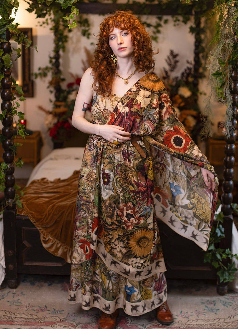 A woman with curly red hair stands in a decorated bedroom wearing a long bohemian floral print maxi dress with wide draped sleeves. The dress features a complex multicolor pattern of flowers, sunflowers, and butterflies on a beige background.