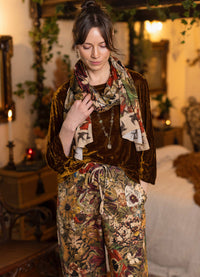 A woman stands in a warmly lit bedroom wearing a dark brown velvet top and matching floral-print pants, with a multicolor botanical scarf draped around her neck.