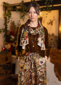 A woman stands indoors wearing a brown velvet long-sleeve top and matching floral-print pants, with a multicolor floral scarf. The setting appears to be a warm, rustic interior with wood furniture and greenery in the background.