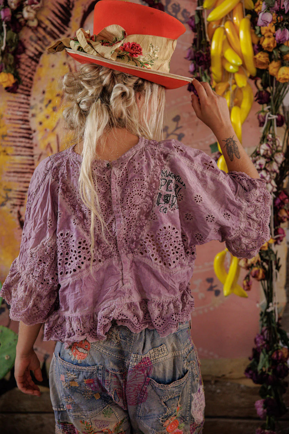 Person wearing a decorative hat and purple lace top in a colorful, artistic setting.