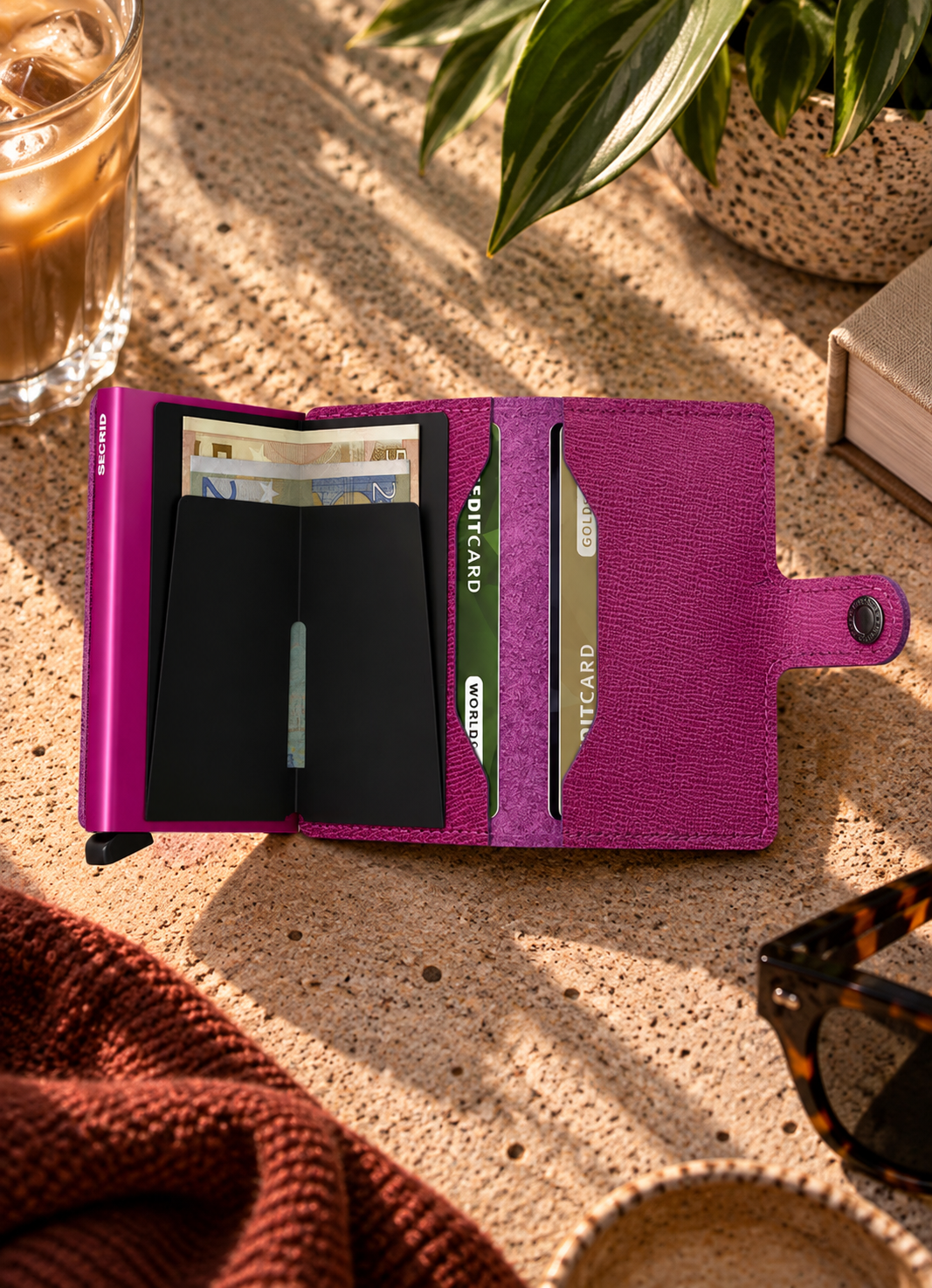 A purple textured leather wallet shown open on a stone surface, with card slots, cash compartments, and a strap closure visible. Surrounding props include an iced coffee, a plant, a book, and sunglasses.