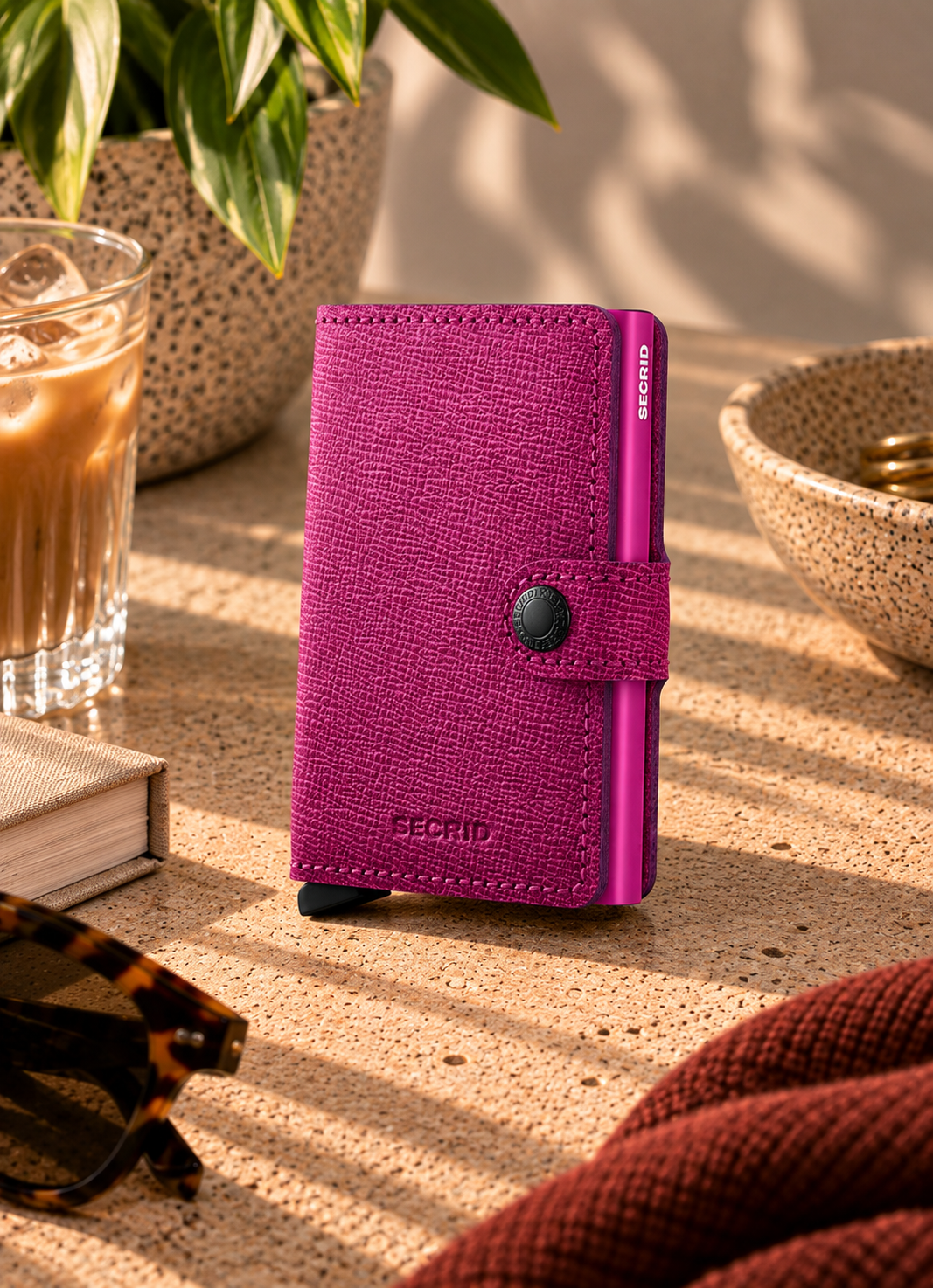 A magenta textured leather wallet with a snap-button strap and debossed brand name, standing on a sunlit tabletop with coffee, glasses, and books nearby.