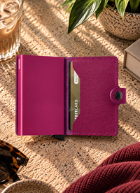 A slim magenta metallic wallet standing upright in a sleeve on a beige stone surface, with a plant, book, and tray in the softly lit background.