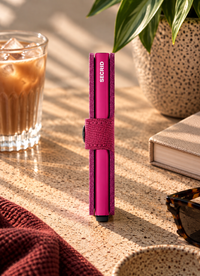 A pink Secrid pen holder standing upright on a textured beige tabletop, surrounded by lifestyle props including an iced coffee, a book, sunglasses, and a potted plant in warm sunlight.