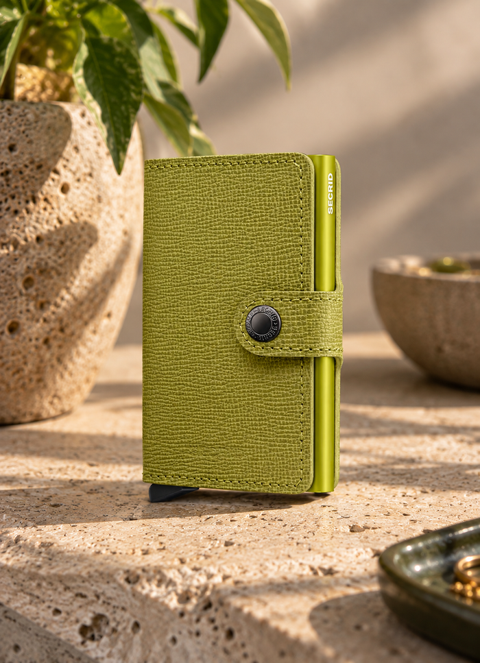 A slim olive green textured wallet standing upright on a stone surface, with a plant and bowls softly blurred in the background.