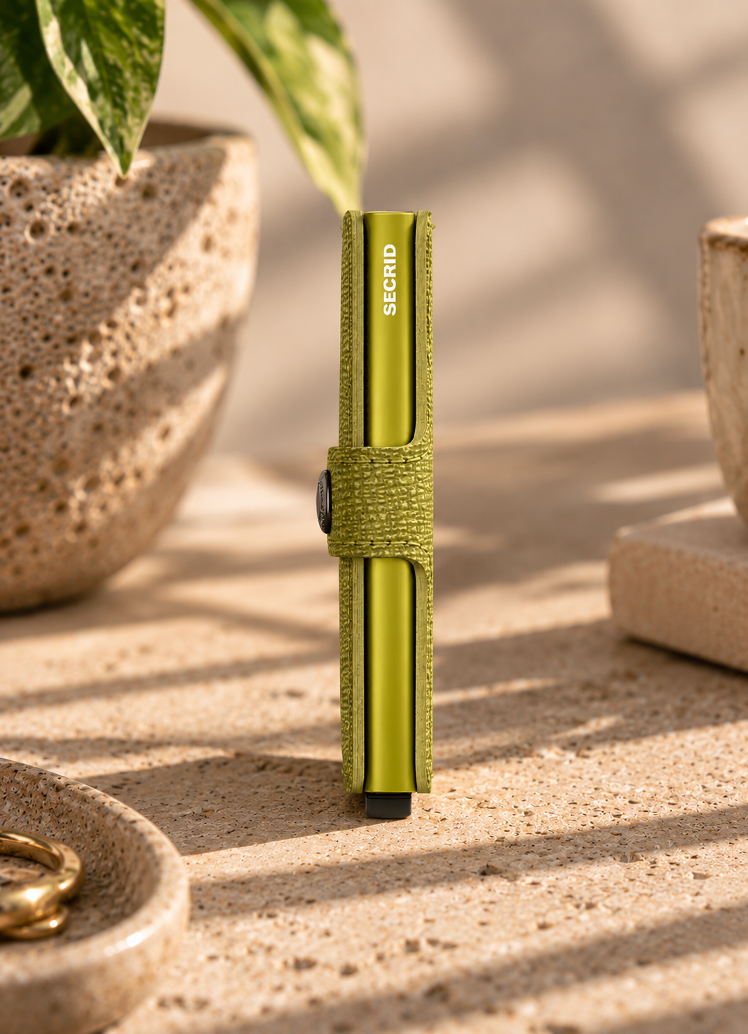 A slim metallic olive-green pen or card holder standing upright on a textured stone surface, wrapped with a matching fabric strap. The word 'SECRIPT' is printed vertically on the front.