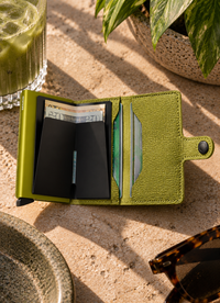An open lime green textured leather wallet with black card sleeves and visible cards, styled on a beige stone surface with a drink, plant leaves, and sunglasses nearby.