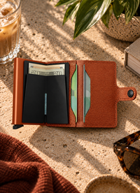 A brown textured leather card holder wallet shown open on a stone surface, with cash, cards, sunglasses, a drink, and a plant nearby in a styled lifestyle setting.