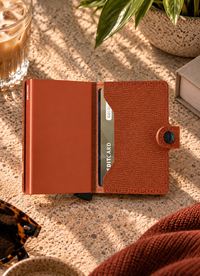 A slim orange metallic wallet laying down  in a textured brown leather sleeve on a beige stone surface, with a plant, book, and tray in the softly lit background.