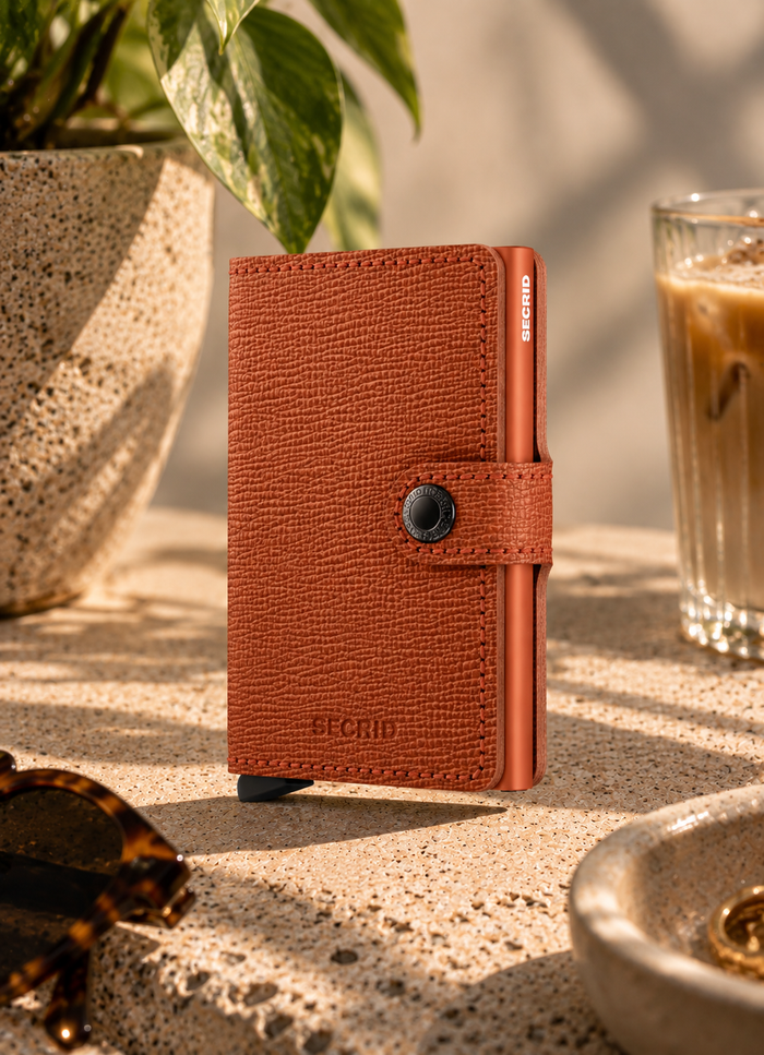 A brown textured leather wallet with a snap-button strap closure, standing upright on a stone surface in a sunlit cafe-like setting with a plant, coffee cup, and sunglasses nearby.