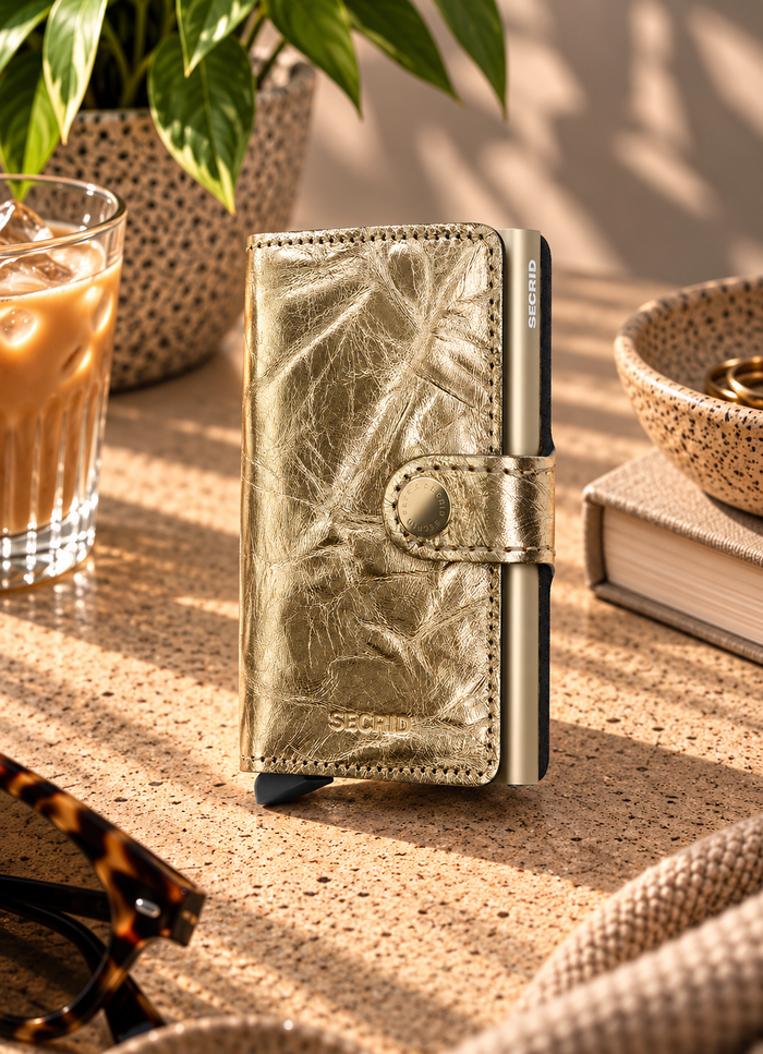 A metallic gold Secured wallet standing upright on a textured beige tabletop, surrounded by lifestyle props including an iced coffee, sunglasses, a book, and a bowl.