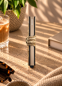 A metallic gold wallet with a crinkled texture and a silver-toned metal interior, shown open on a textured beige surface with lifestyle props including sunglasses, a drink, and a plant.