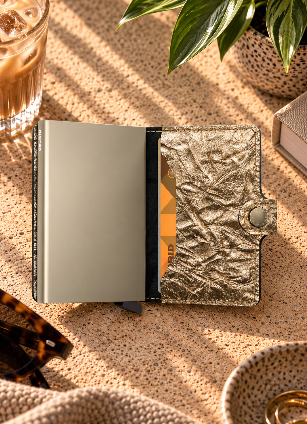 A metallic gold wallet with a crinkled texture and a silver-toned metal interior, shown open on a textured beige surface with lifestyle props including sunglasses, a drink, and a plant.