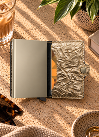 A metallic gold wallet with a crinkled texture and a silver-toned metal interior, shown open on a textured beige surface with lifestyle props including sunglasses, a drink, and a plant.