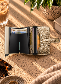 A metallic gold minimalist wallet with black interior card slots, shown open on a textured beige surface with warm sunlight and surrounding lifestyle props.